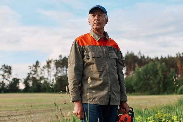 Portrait of mature man wearing jacket and cap posing outdoor in meadow with chainsaw in hands