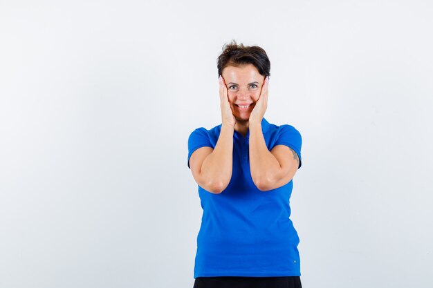 Portrait of mature woman holding hands on cheeks in blue t-shirt and looking merry front view