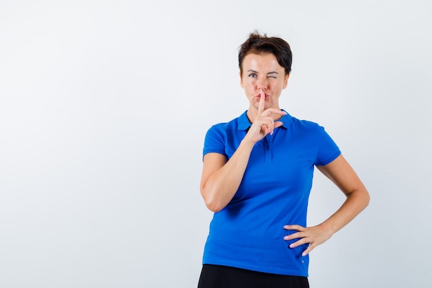 Портрет зрелой женщины, показывающей жест тишины, подмигивающей глазом в синей футболке и уверенно выглядящей, вид спереди
