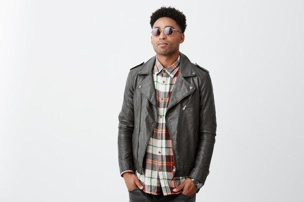 Portrait of mean good-looking dark-skinned guy with afro hairstyle in checkered shirt under leather jacket and sunglasses holding hands in pockets, waiting for girlfriend outside cafe.