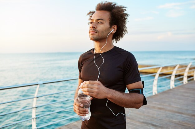 Портрет медлительного и сосредоточенного темнокожего спортсмена с густыми волосами, держащего в руках бутылку минеральной воды.