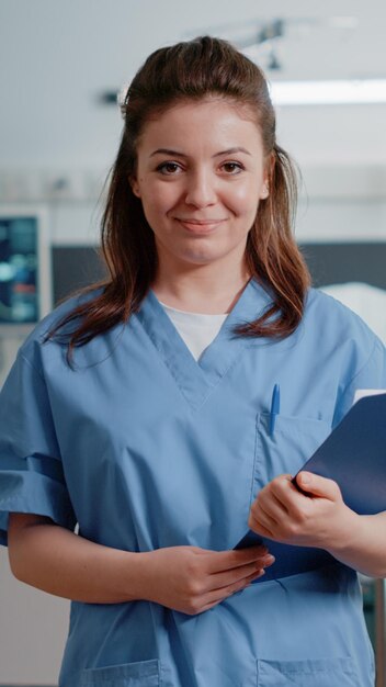 Portrait of medical assistant holding checkup documents to help doctor with treatment and medicine. Woman working as nurse with uniform and tools looking at camera to cure patient