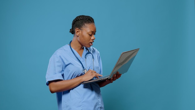 医療システムに取り組んで医療を実践するためにラップトップディスプレイを見ている制服を着た医療看護師の肖像画。専門知識を持つためにコンピューターを使用して聴診器を持つ女性アシスタント。