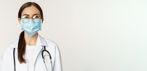 Ritratto di medico donna operatore medico in maschera facciale da covid durante la pandemia sorridente e guardando entusiasta in piedi su sfondo bianco