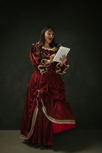 Portrait of medieval young woman in red vintage clothing using tablet on dark background.