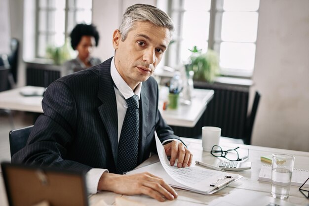 Portrait of mid adult businessman sitting at desk and going through business reports while looking at camera in the office