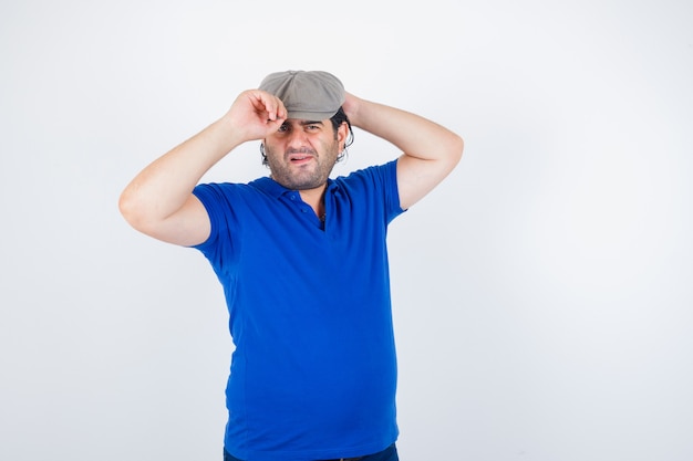Portrait of middle aged man adjusting his cap in polo t-shirt, ivy hat and looking indecisive front view