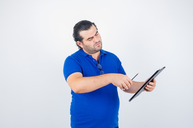 Portrait of middle aged man looking through clipboard while holding pencil in polo t-shirt and looking thoughtful front view