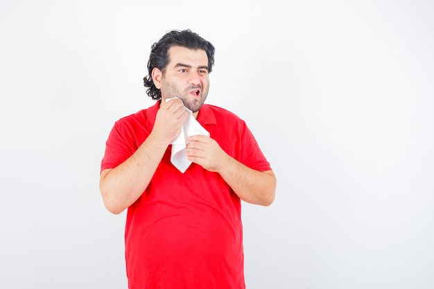 Ritratto di uomo di mezza età asciugandosi la guancia con un tovagliolo in maglietta rossa e guardando premurosa vista frontale