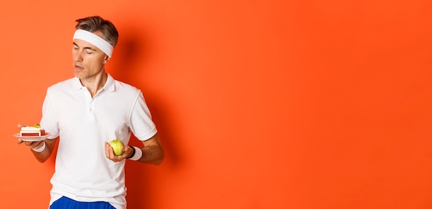 Free photo portrait of middleaged sportsman making decision between green apple and cake being on diet standing