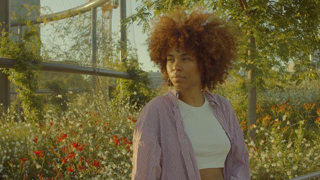 Портрет чернокожей женщины смешанной расы в парке, полном цветов, с огромными афро-волосами в контурном солнечном свете, смотрящей в сторону цвета кинопленки