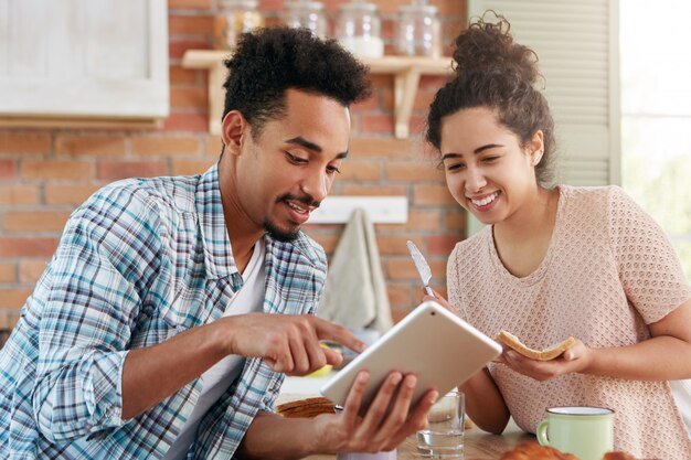 混合レースの流行に敏感な男の肖像画は、サンドウィッチの作成で忙しい妻にタブレットコンピューターで何かを示しています