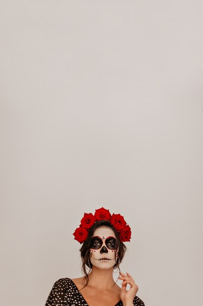 Portrait of model against white wall, posing in crown of natural flowers. Halloween skeleton makeup looks unusual