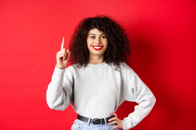 Portrait of modern european woman with curly hair showing number one, making an order, raising finger and smiling, standing on red background.