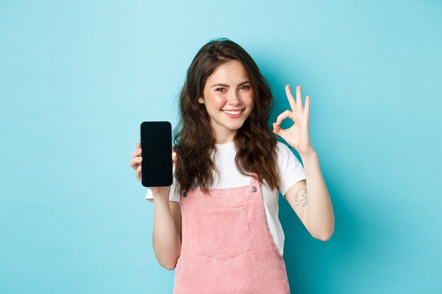 Портрет современной стильной девушки, рекомендующей интернет-магазин или мобильное приложение, показывающий знак «хорошо» с пустым экраном смартфона, одобрительный кивок, удовлетворенная улыбка, синий фон.