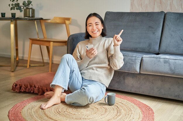 portrait of modern young korean woman sitting on floor pointing finger at copy space showing banner