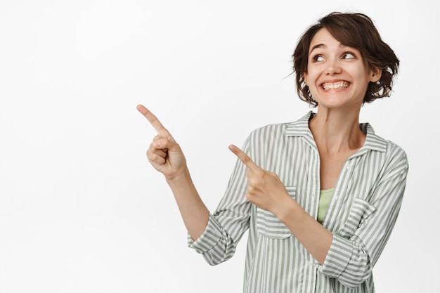 Free photo portrait of modern young woman pointing fingers left, looking excited standing on white.