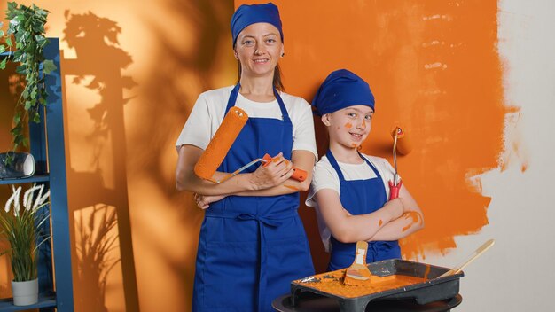 Portrait of mom and child painting walls with orange color paint, using renovating equipment and roller brush tools to redecorate apartment interior. Home decor improvement housework.