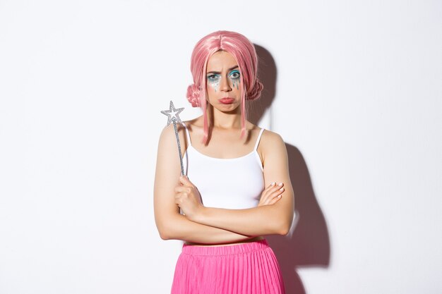 Portrait of moody cute girl in pink wig, dressed as a fairy for halloween, looking upset or disappointed, standing over white background