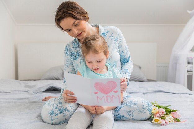 Portrait of mother and daughter reading greeting card on bed at home