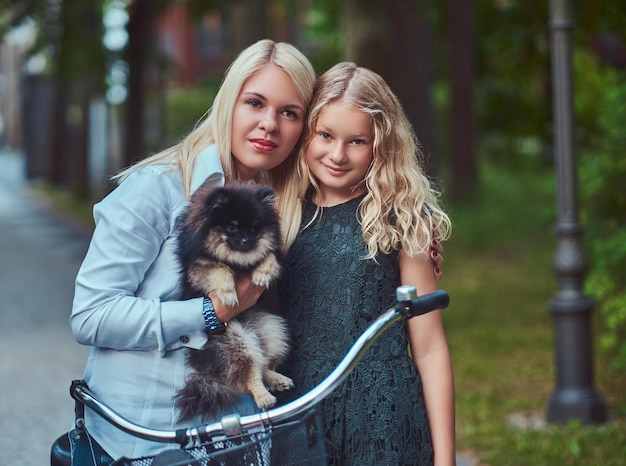 Foto gratuita ritratto di una madre e una figlia con i capelli biondi su un giro in bicicletta con il loro simpatico cagnolino spitz in un parco.