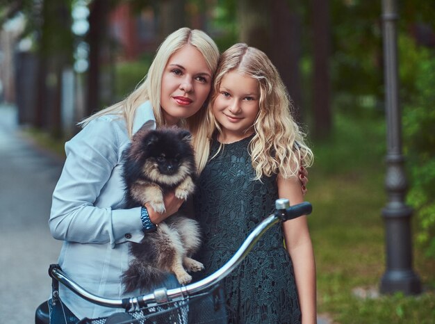 Portrait of a mother and daughter with a blonde hair on a bicycle ride with their cute little spitz dog in a park.