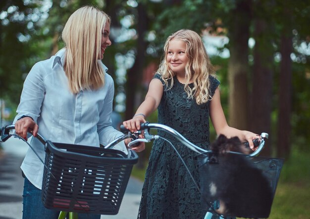 공원에서 귀여운 작은 스피츠 개와 함께 자전거를 타고 있는 금발 머리를 한 엄마와 딸의 초상화.