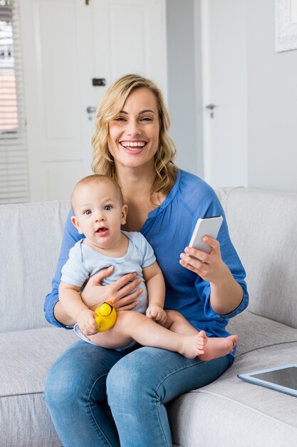 Portrait of mother holding baby boy and using mobile phone in living room