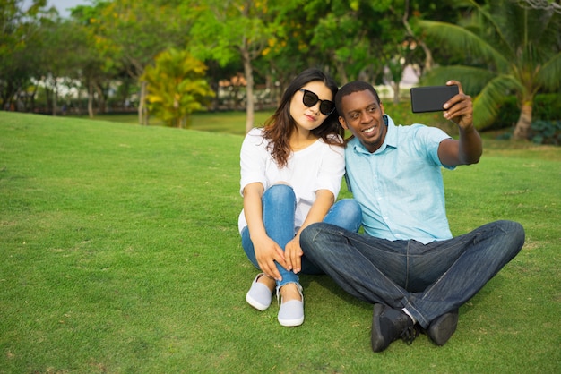 Portrait of multiethnic couple siting on lawn and taking selfie with smartphone.