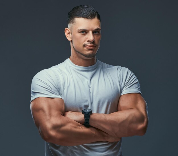スタジオで腕を組んで立っている、スポーツウェアの筋肉質のハンサムなボディービルダーの肖像画。灰色の背景に分離。