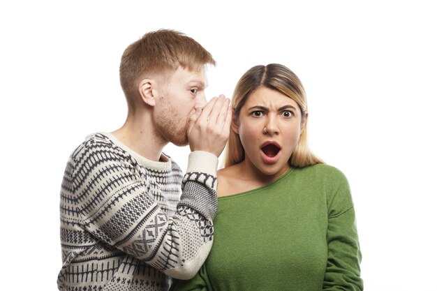 Portrait of mysterious young fair-haired male with stubble covering ear of his shocked girlfriend who is listening to gossips, rumours, confidential information and secrets with eyes full of disbelief