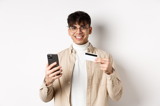 Portrait of natural young man in glasses paying in internet, showing smartphone and plastic credit card, standing on white background