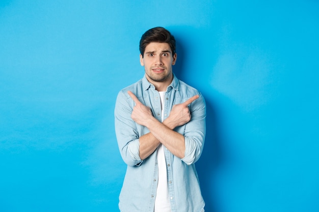 Portrait of nervous guy pointing fingers sideways, looking indecisive and asking for help with choice, showing left and right promo offers, standing against blue background