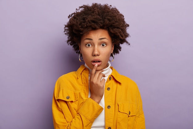 Портрет нервной, озадаченной молодой женщины касается нижней губы, у нее кудрявая афро-прическа, тревожное выражение лица, в модной желтой куртке и сережках, модели в помещении на фиолетовом фоне