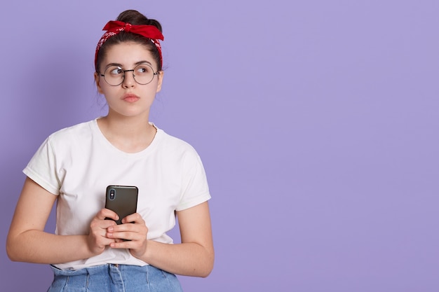 Portrait of nice attractive girl holding cell in hands looking aside with pensive expression isolated over lilac space