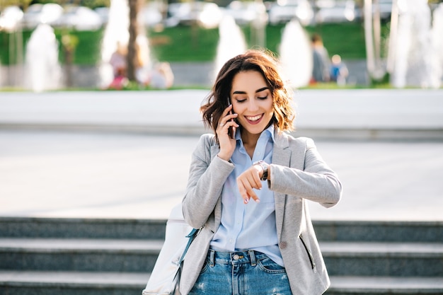 Ritratto di una bella ragazza bruna con i capelli corti che cammina in città. indossa camicia, jeans, giacca e borsa. sta parlando al telefono, guardando l'orologio e sorridendo.