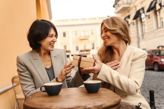 Portrait of nice caucasian adult lady congratulates her stylish girlfriend by giving gift in beige package with brown bow Rest leisure concept