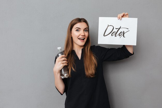 Portrait of nice smiling lady standing and holding postcard and bottle of water while happily looking in camera on gray background