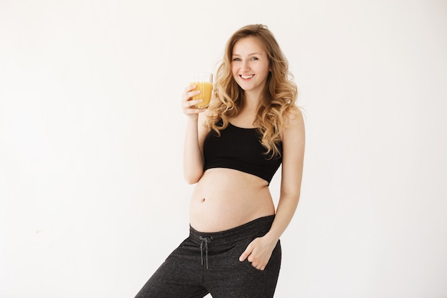 Portrait o young good-looking joyful pregnant woman with light hair in comfy home clothes smiling, holding glass of orange juice in hand, ready to start her day with tasty drink.