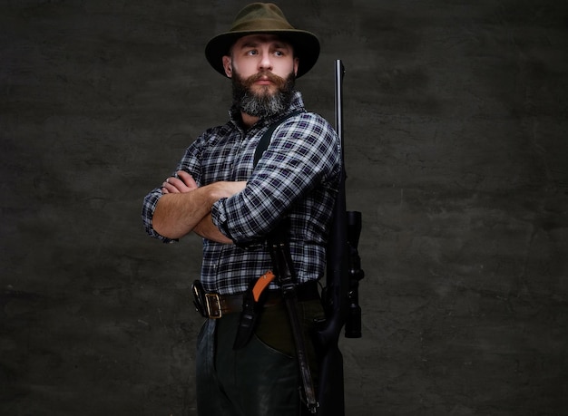 Бесплатное фото Портрет бородатого охотника в флисовой рубахе и шапке, стоящего с винтовкой за спиной. изолированные на темном фоне.