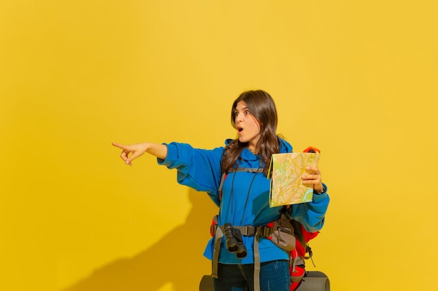 黄色​の​スタジオ​の​壁​に​分離された​バッグ​と​双眼鏡​で​陽気な​若い​観光​の​女の子​の​肖像画