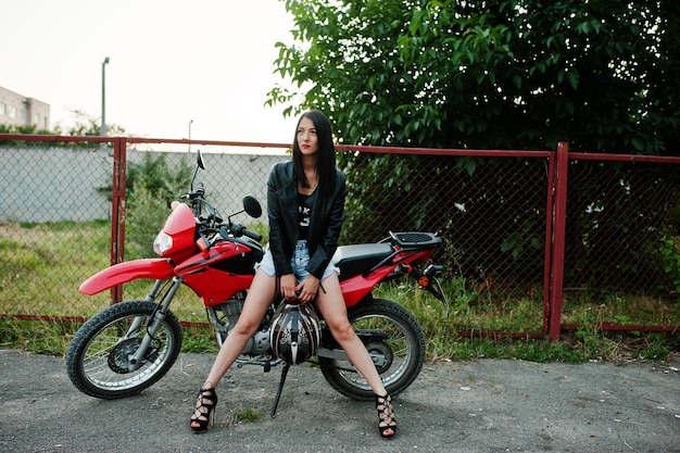 Бесплатное фото Портрет крутой и потрясающей женщины в платье и черной кожаной куртке, сидящей на крутом красном мотоцикле