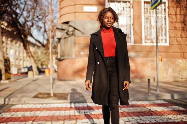 無料写真 横断歩道を歩いているファッショナブルな黒いコートと赤いタートルネックを身に着けている縮れ毛のアフリカの女性の肖像画