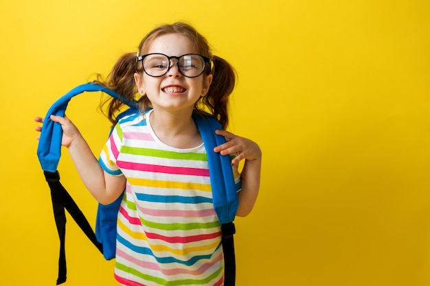 Портрет девушки в очках и полосатой футболке со школьным рюкзаком на желтом фоне. радостный ребенок спешит в школу. концепция образования. фотостудия, место для текста.