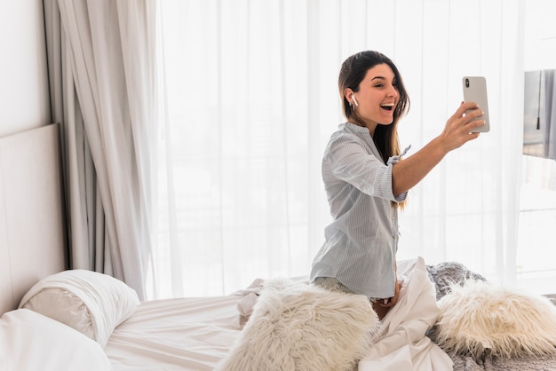 携帯​電話​で​ビデオ​通話​を​取って​ベッド​の​上に​座って​幸せな​若い​女​の​肖像
