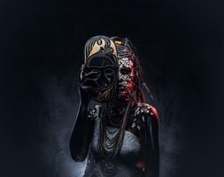 portrait of a scary african shaman female with a petrified cracked skin and dreadlocks, holds a traditional mask on a dark background. make-up concept.