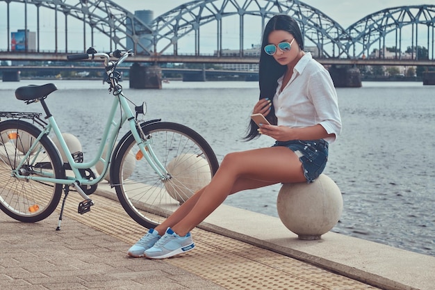 Бесплатное фото Портрет сексуальной брюнетки, одетой в блузку и шорты в солнцезащитных очках, сидящей на набережной у моста, использующей смартфон, расслабляющейся после езды на велосипеде.