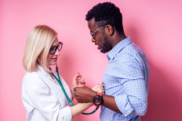 アフロアメリカ​の​患者​の​男性​と​笑顔​の​女性​医師​の​肖像画​。​美しい​ブロンド​の​女性​の​白い​医療​コート​と​スタジオ​の​ピンク​の​背景​に​病気​の​心臓​に​聴診器​を​保持している​メガネ​。