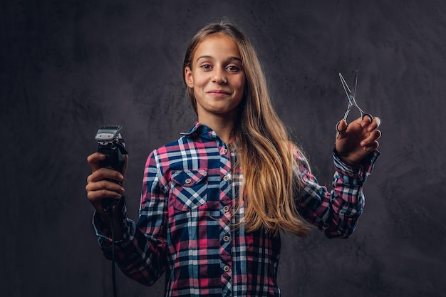 Бесплатное фото Портрет улыбающейся молодой девушки, одетой в рубашку, с триммером и ножницами. изолированные на темном текстурированном фоне.