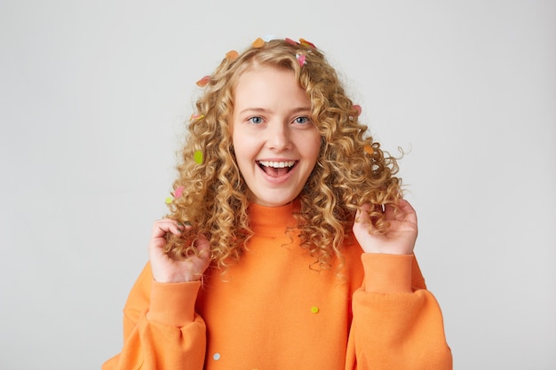Портрет очень счастливой девушки в оранжевом свитере играет с вьющимися волосами, улыбаясь изолированной на белой стене